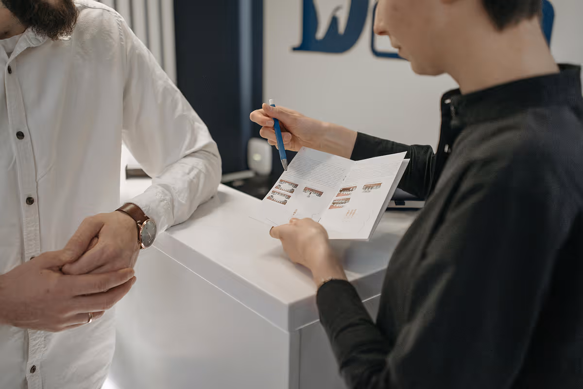 Person in black shirt holds booklet and pen, showing pages to a person in white shirt at a counter.