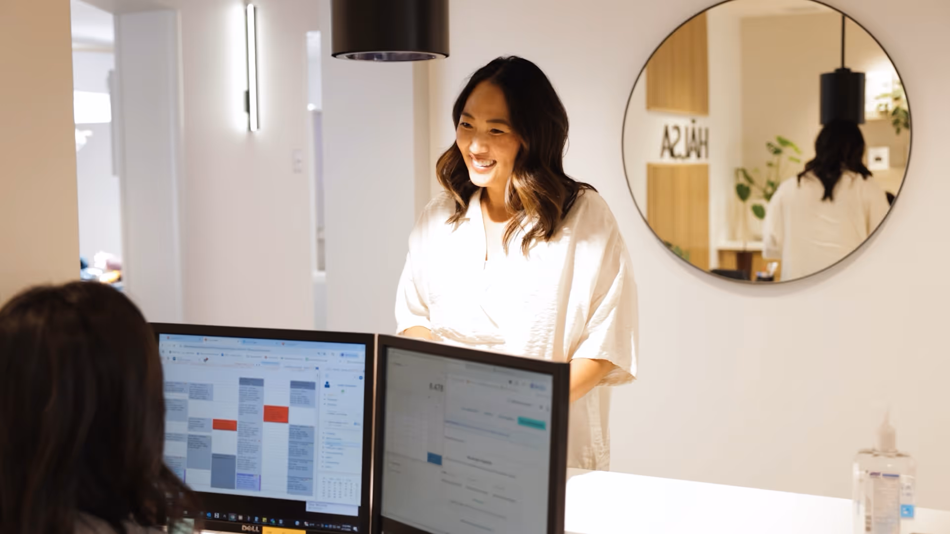 Smiling woman standing and talking to colleague seated at desk with dual computer monitors.