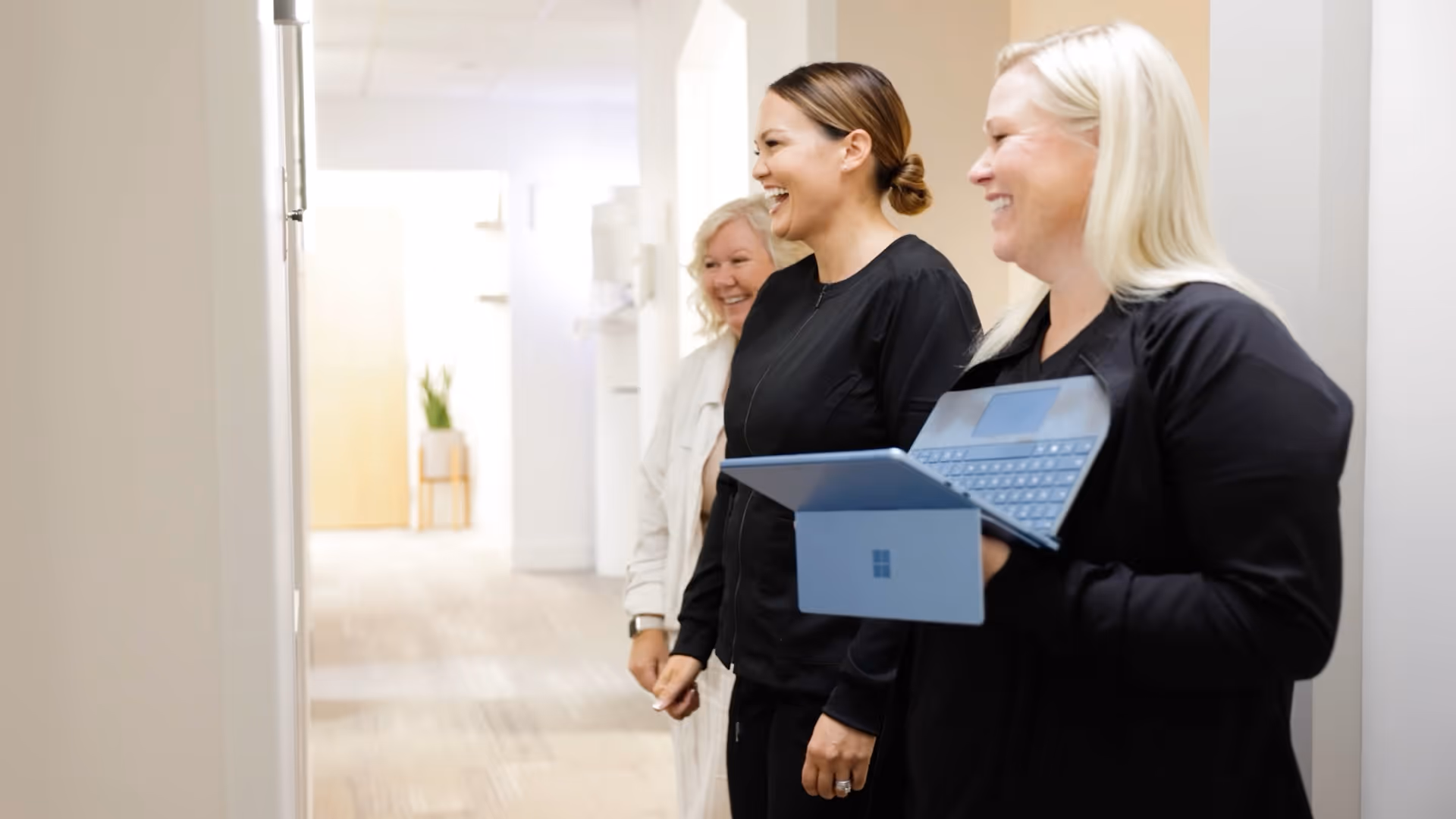 Three women smiling and standing in a hallway, one holding a blue laptop.