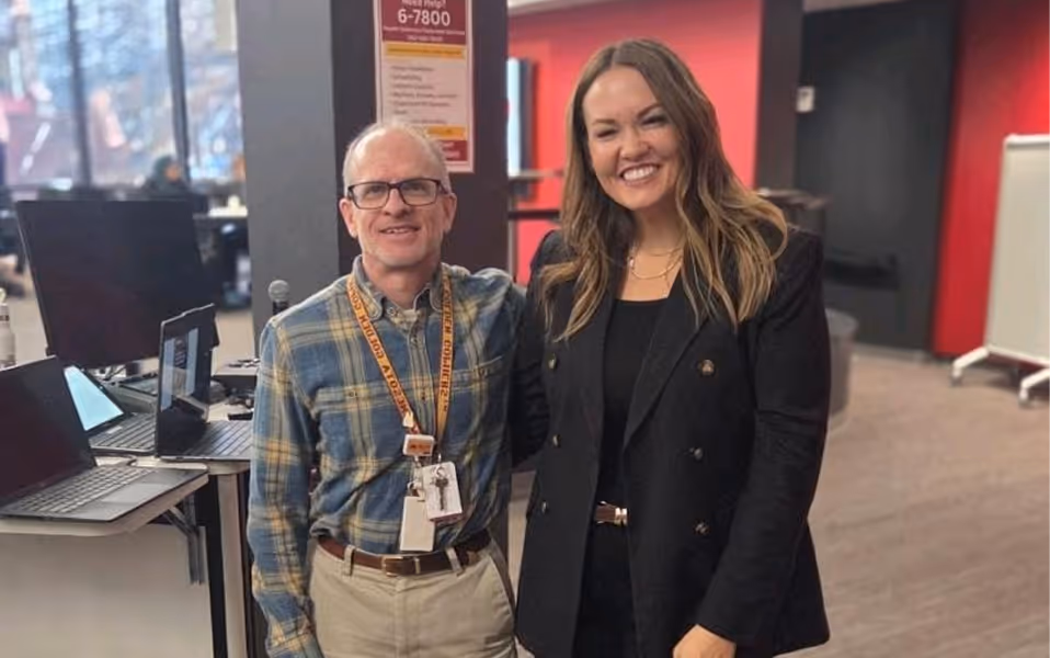 Smiling man in plaid shirt and woman in black blazer posing together indoors near computers.