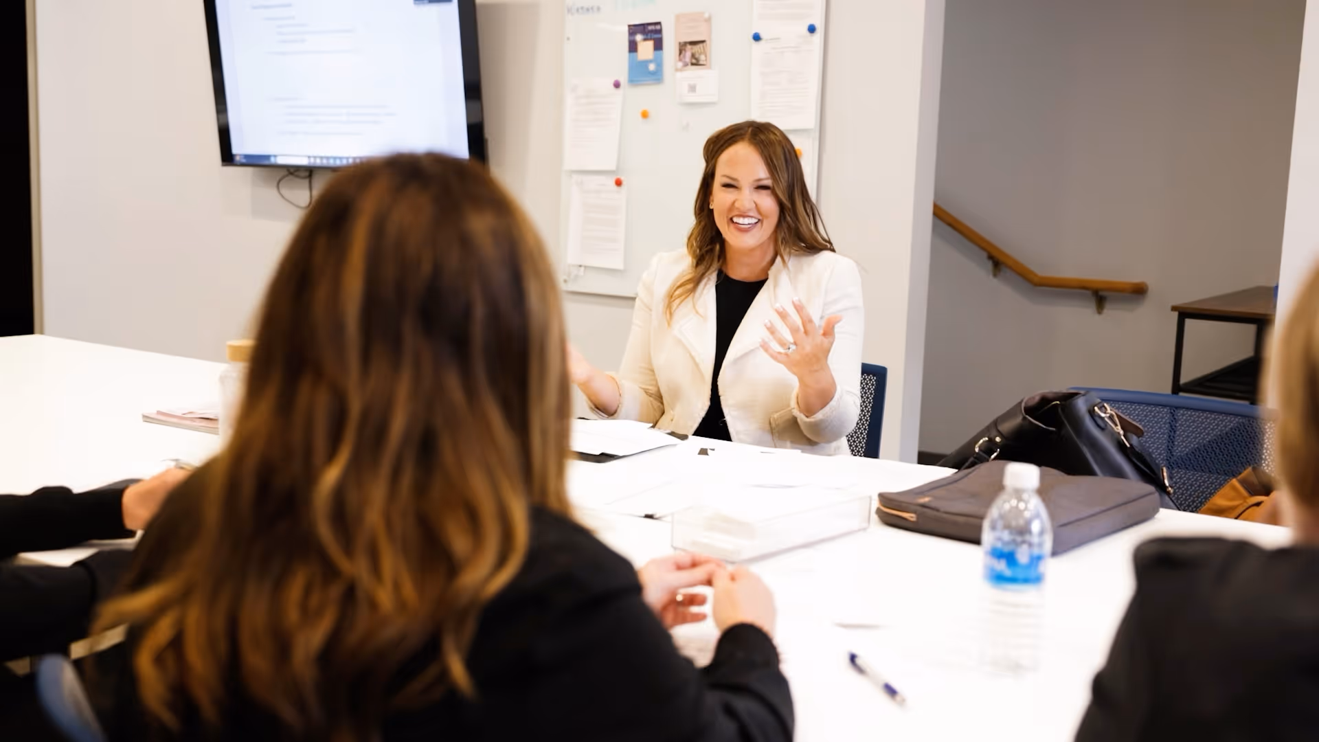 Smiling woman in white jacket speaking to people around a conference table with documents and water bottle.