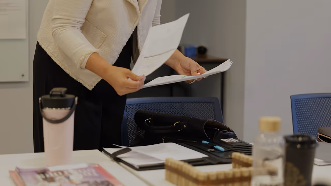 Person sorting papers at a cluttered office desk with notebooks, pens, cups, and bags around.