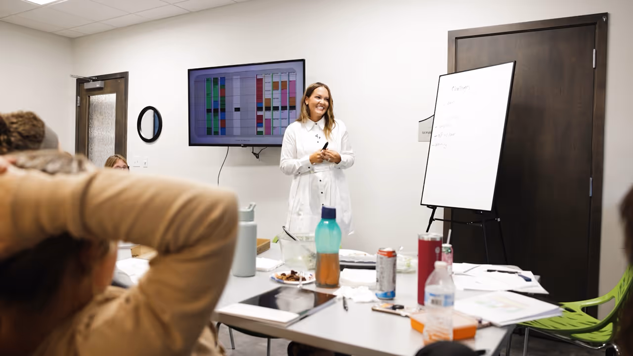 Woman in white shirt leading a meeting with a whiteboard and screen showing colorful charts.