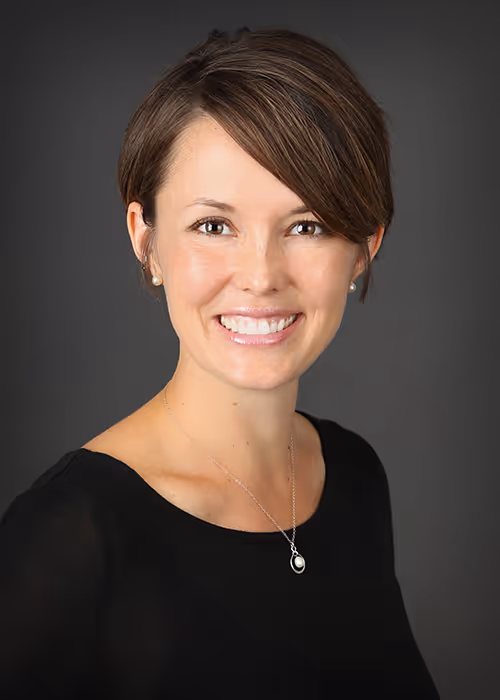 Smiling woman with short brown hair wearing a black top and silver necklace on a dark background.