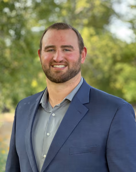 Smiling man with beard wearing a blue suit and light gray shirt outdoors with trees in the background.