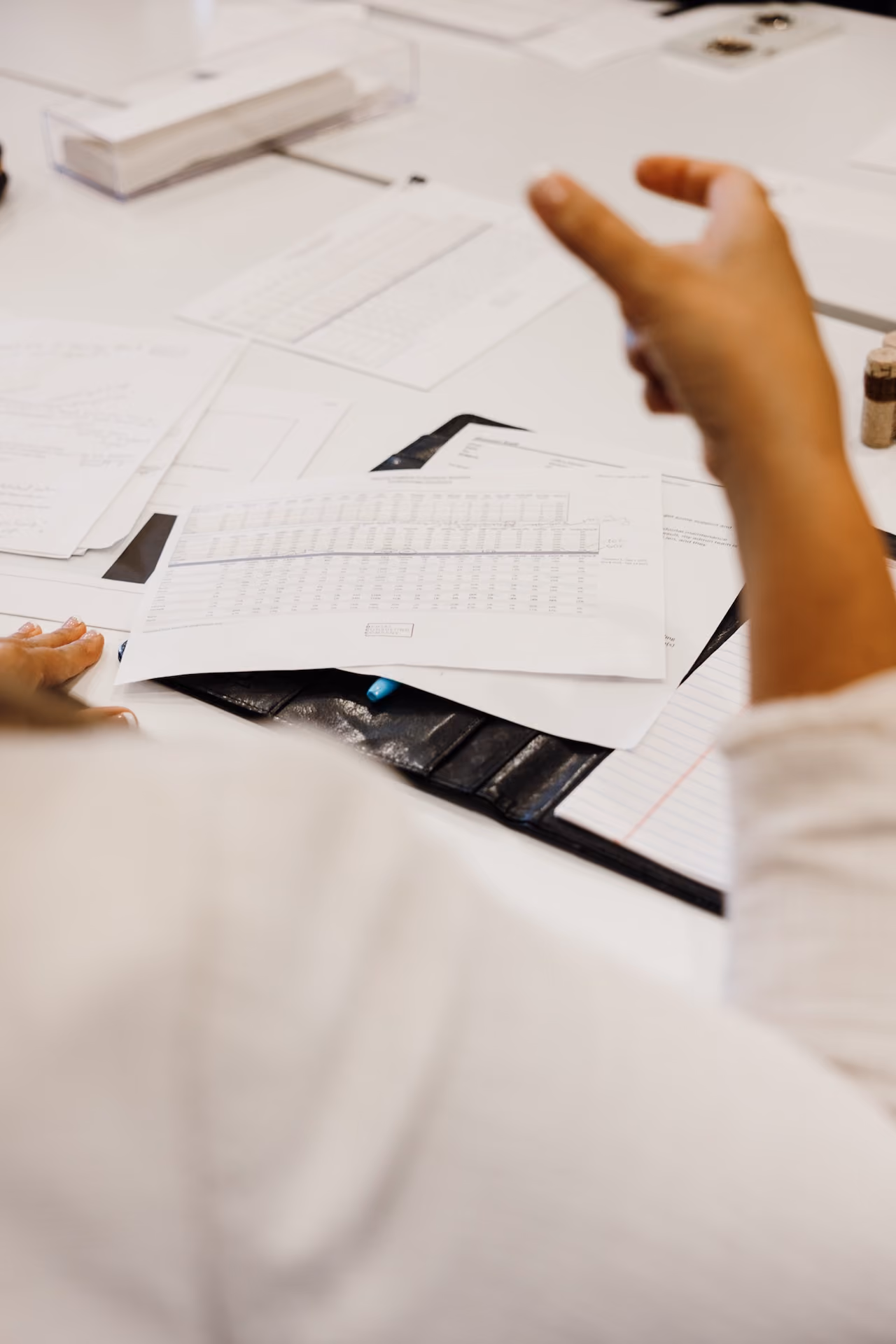 Person gesturing over a white desk with scattered papers, notebooks, and a pen.