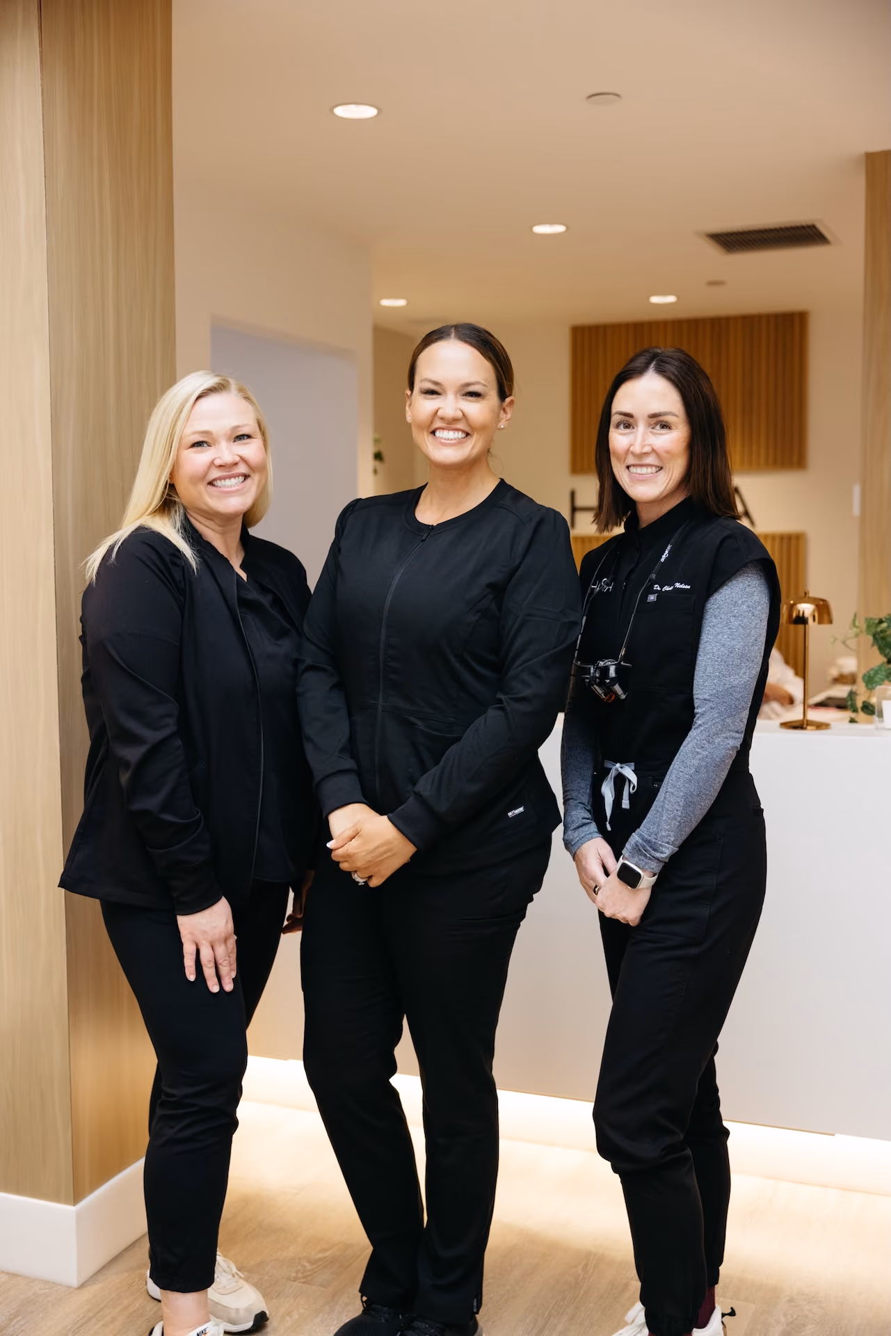 Three women in black medical scrubs smiling inside a modern clinic reception area.
