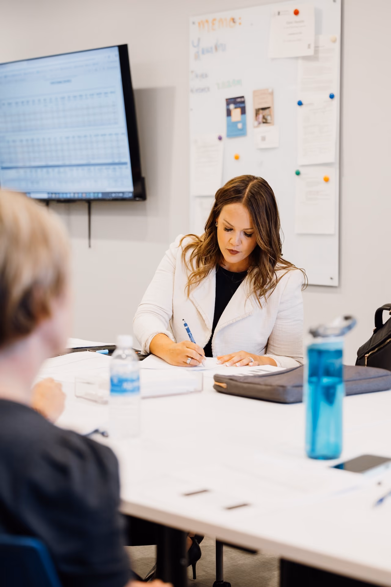 Woman in white blazer writing on paper at a conference table with another person and screen in background.