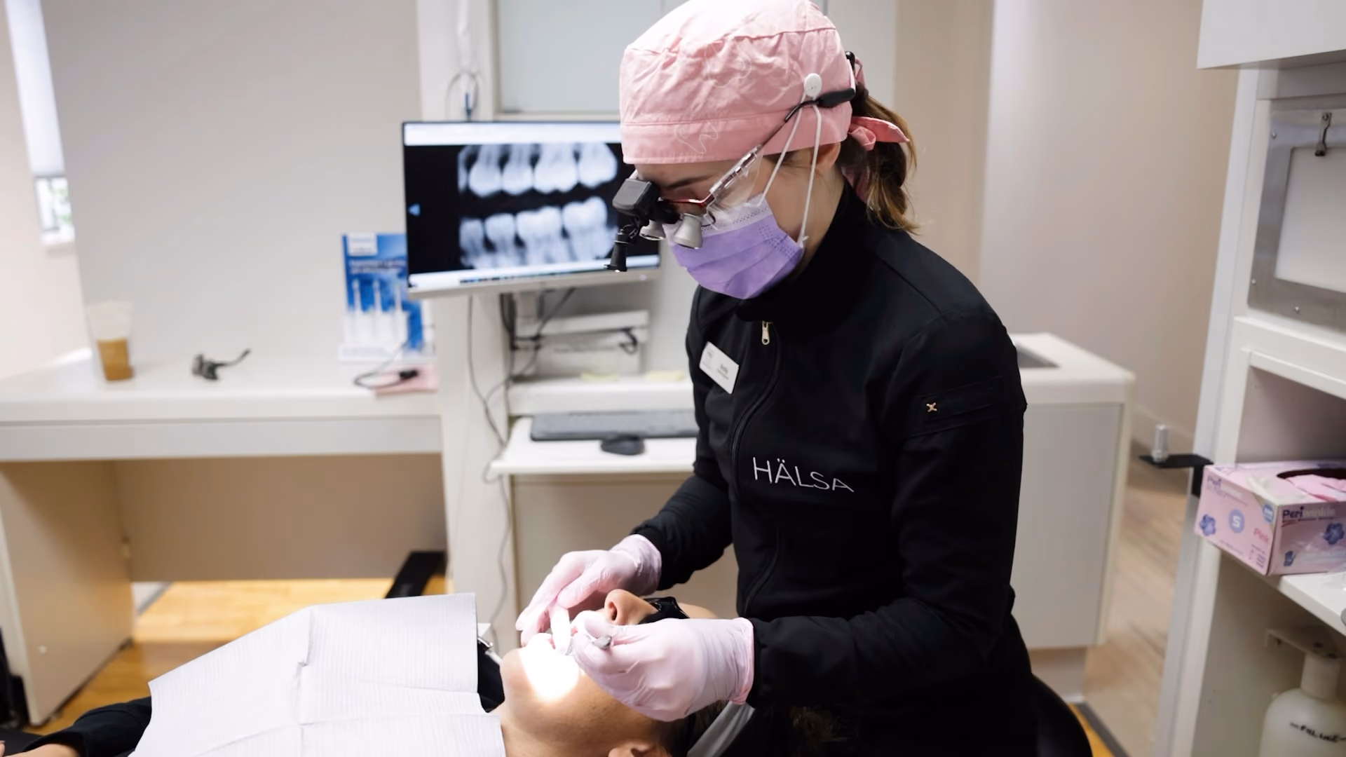Dentist in pink cap and purple mask examining patient's mouth with dental tools in clinic.