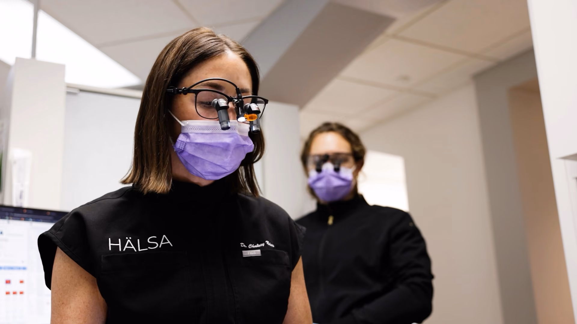 Two medical professionals wearing black, protective glasses, and purple face masks in a clinical setting.