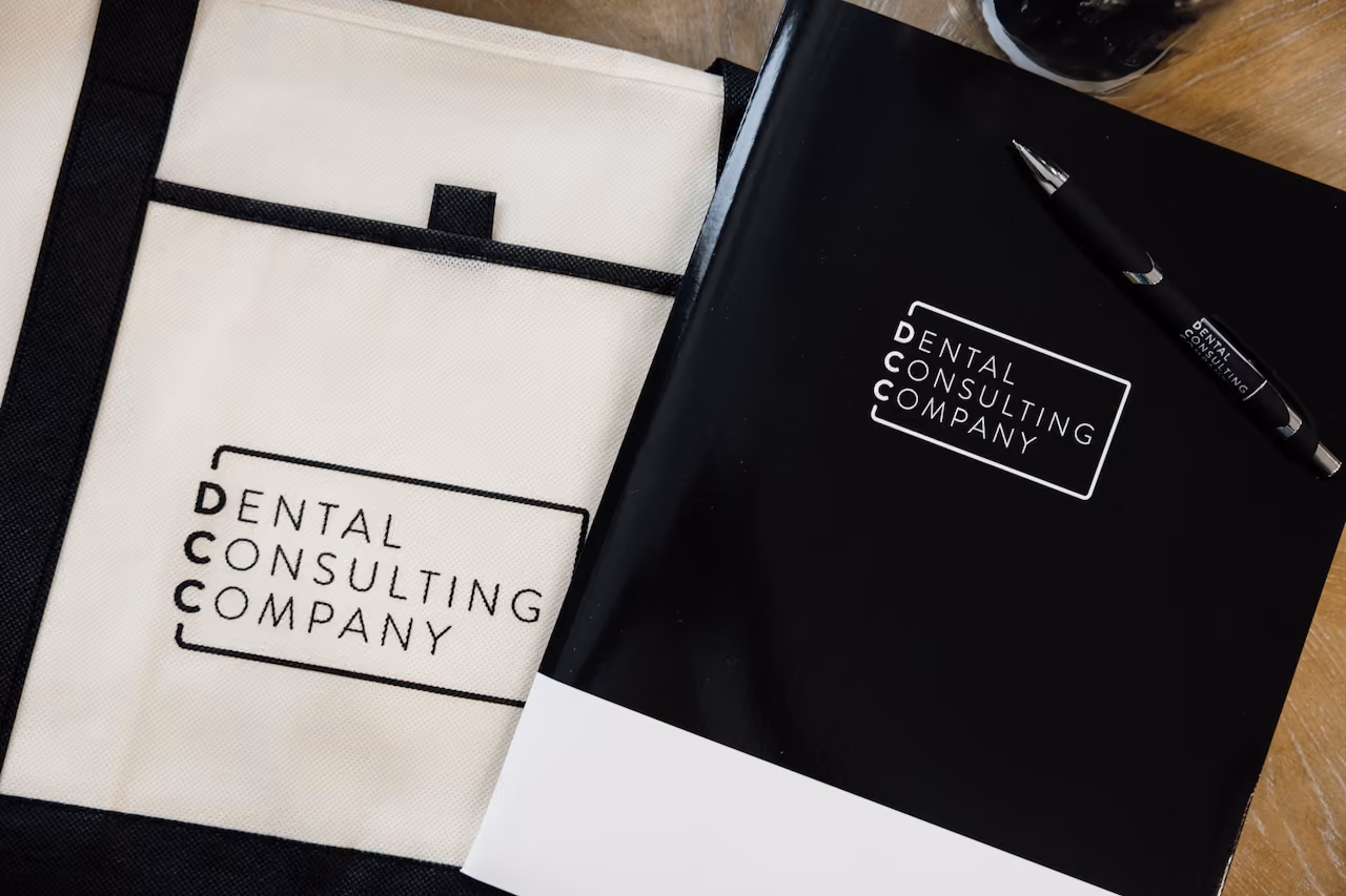 Black and white company materials with logo reading Dental Consulting Company on a folder, pen, and bag.