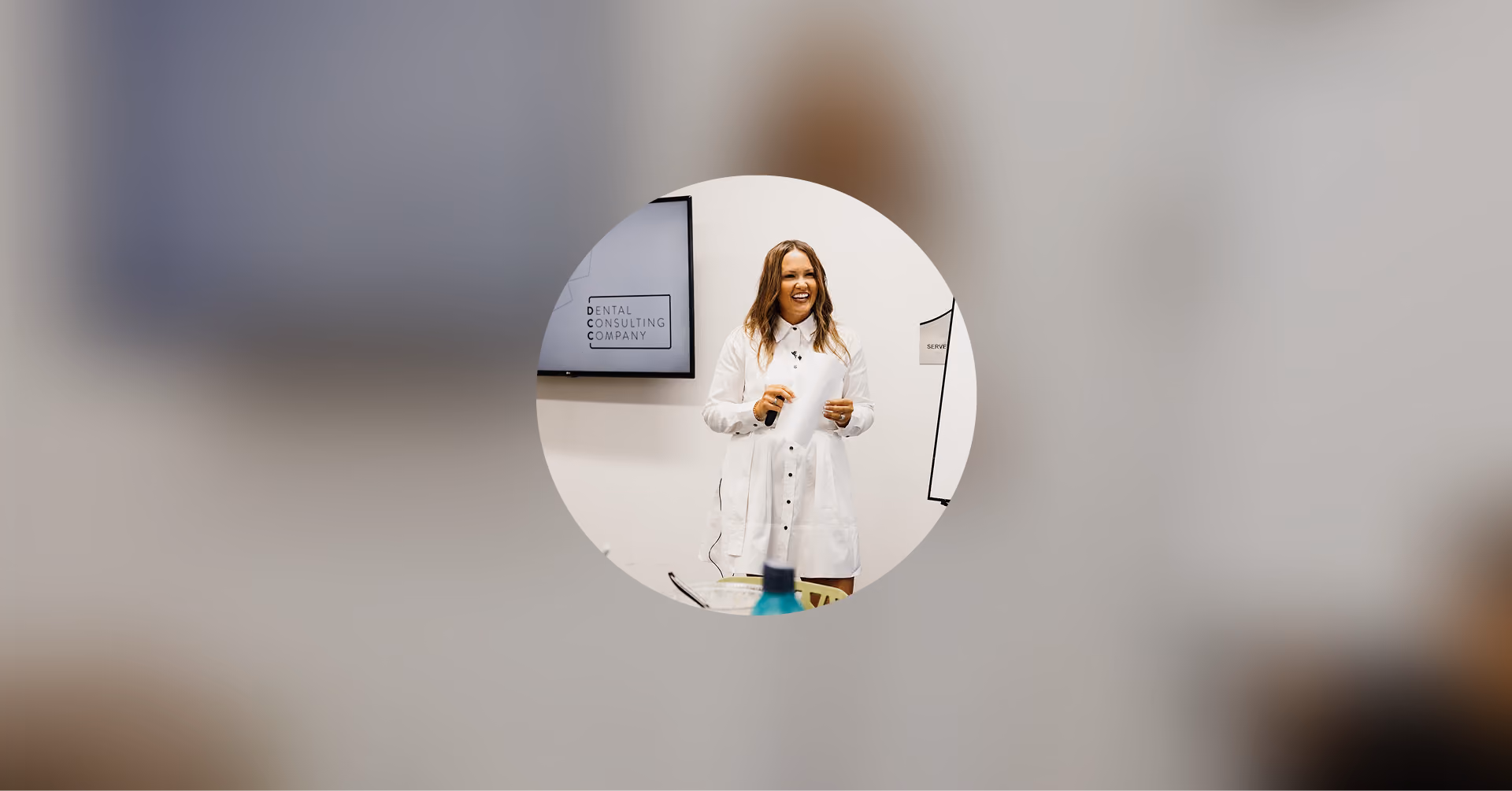 Woman in white shirt dress smiling and holding a remote, standing by a screen labeled Dental Consulting Company.