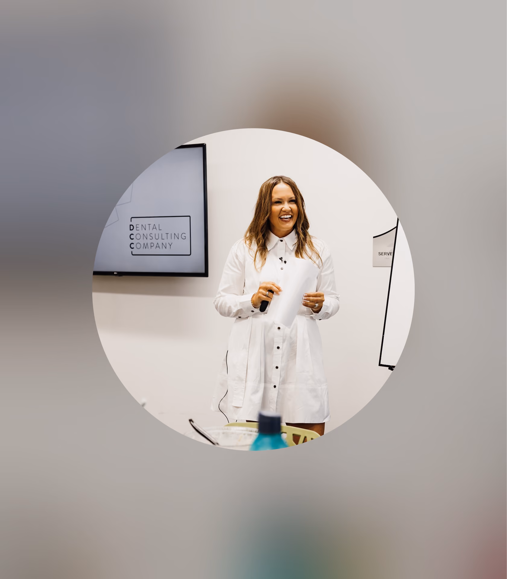 Woman in white shirt dress smiling and holding a remote, standing by a screen labeled Dental Consulting Company.