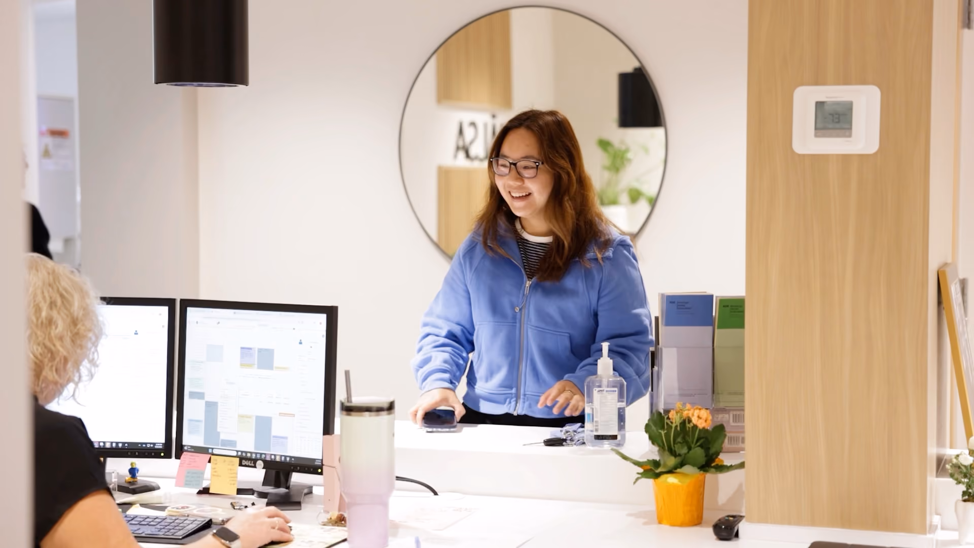 Smiling woman in blue jacket stands at a reception desk with sanitizer and plant nearby.