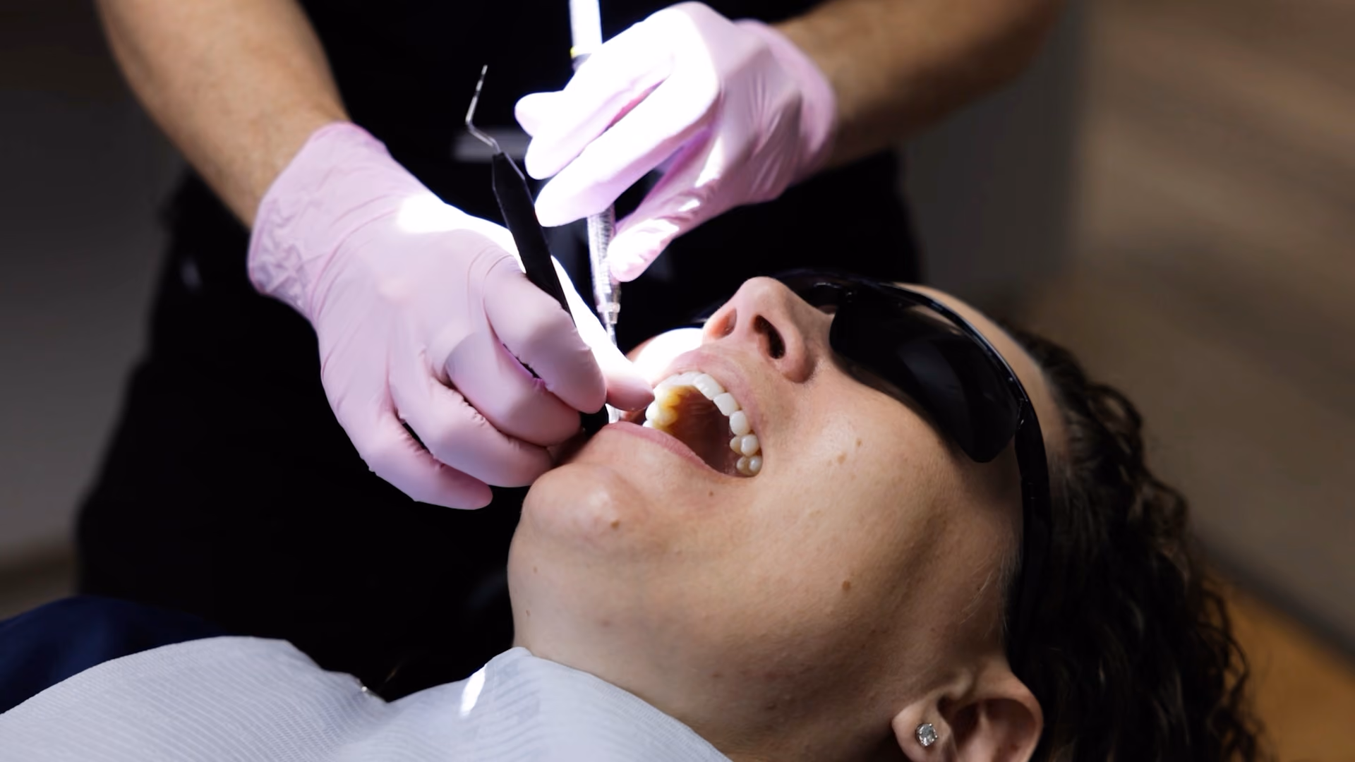 Dentist wearing pink gloves performing dental procedure on patient with sunglasses in dental chair.