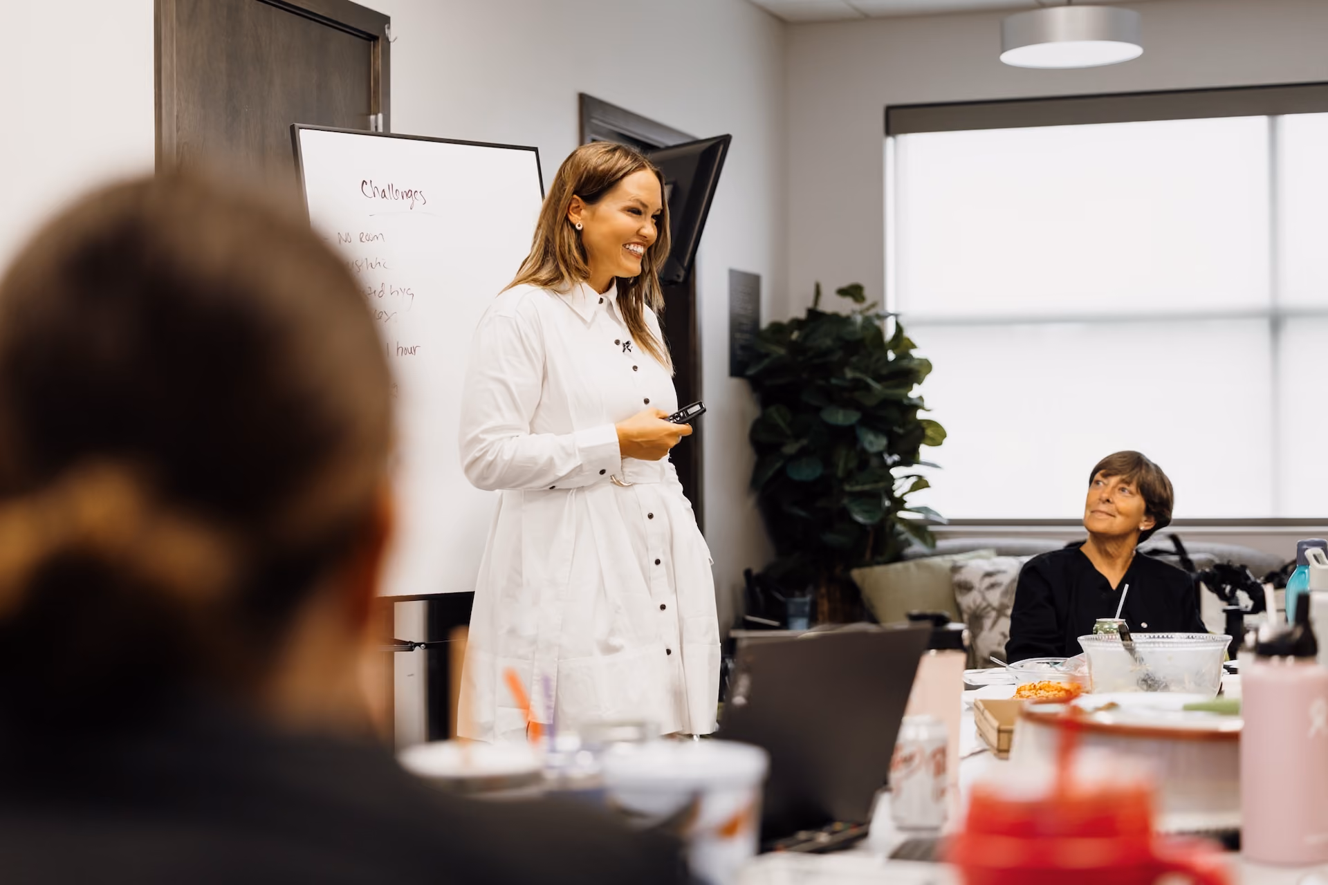 Woman in white shirt presenting by a whiteboard to seated colleagues in a meeting room.