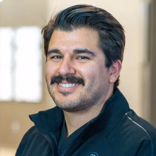 Smiling man with dark hair and mustache wearing a black zip-up jacket indoors.