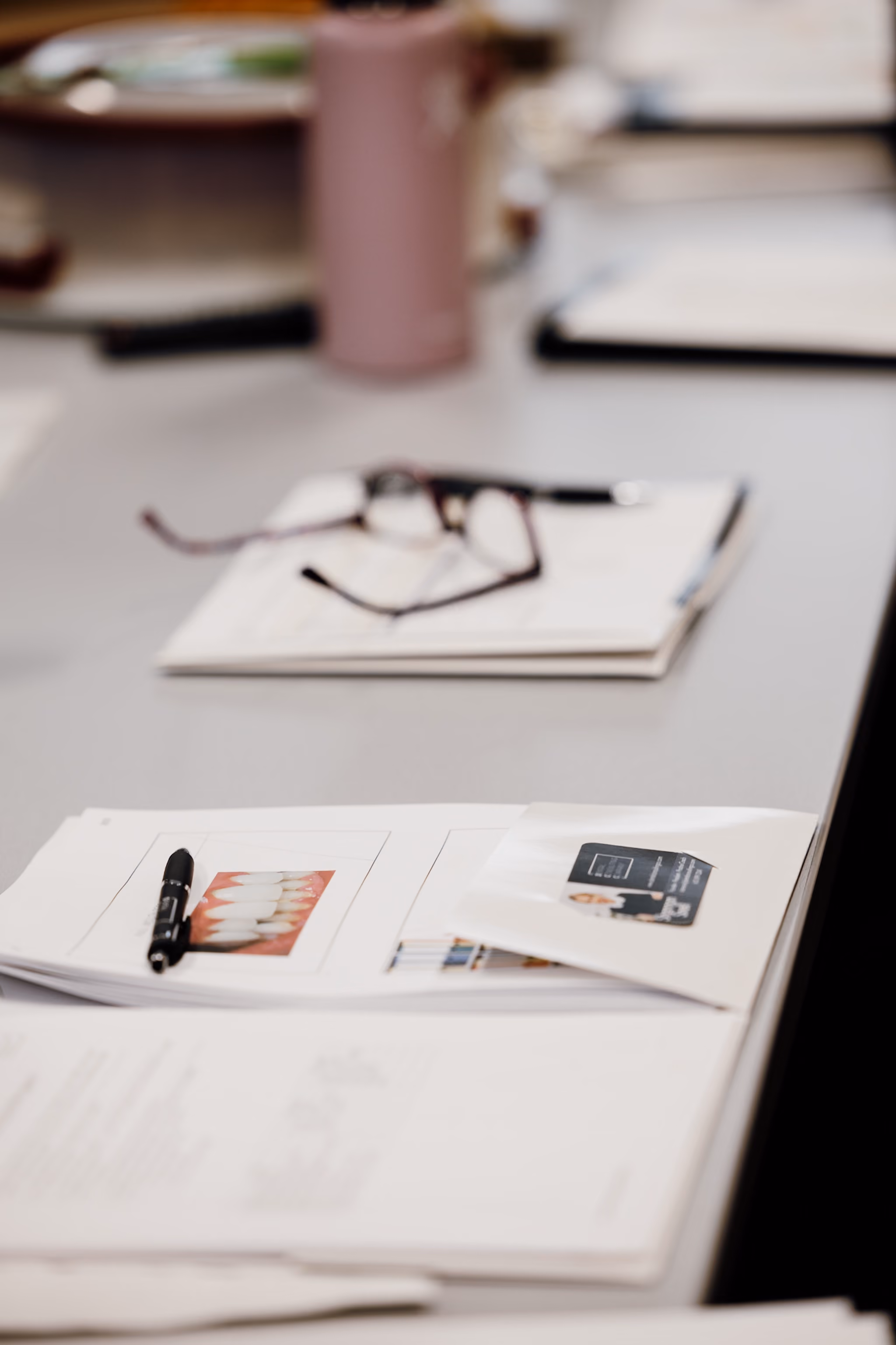 Desk with scattered papers, a pen, eyeglasses, and a pink cup in a blurred background.