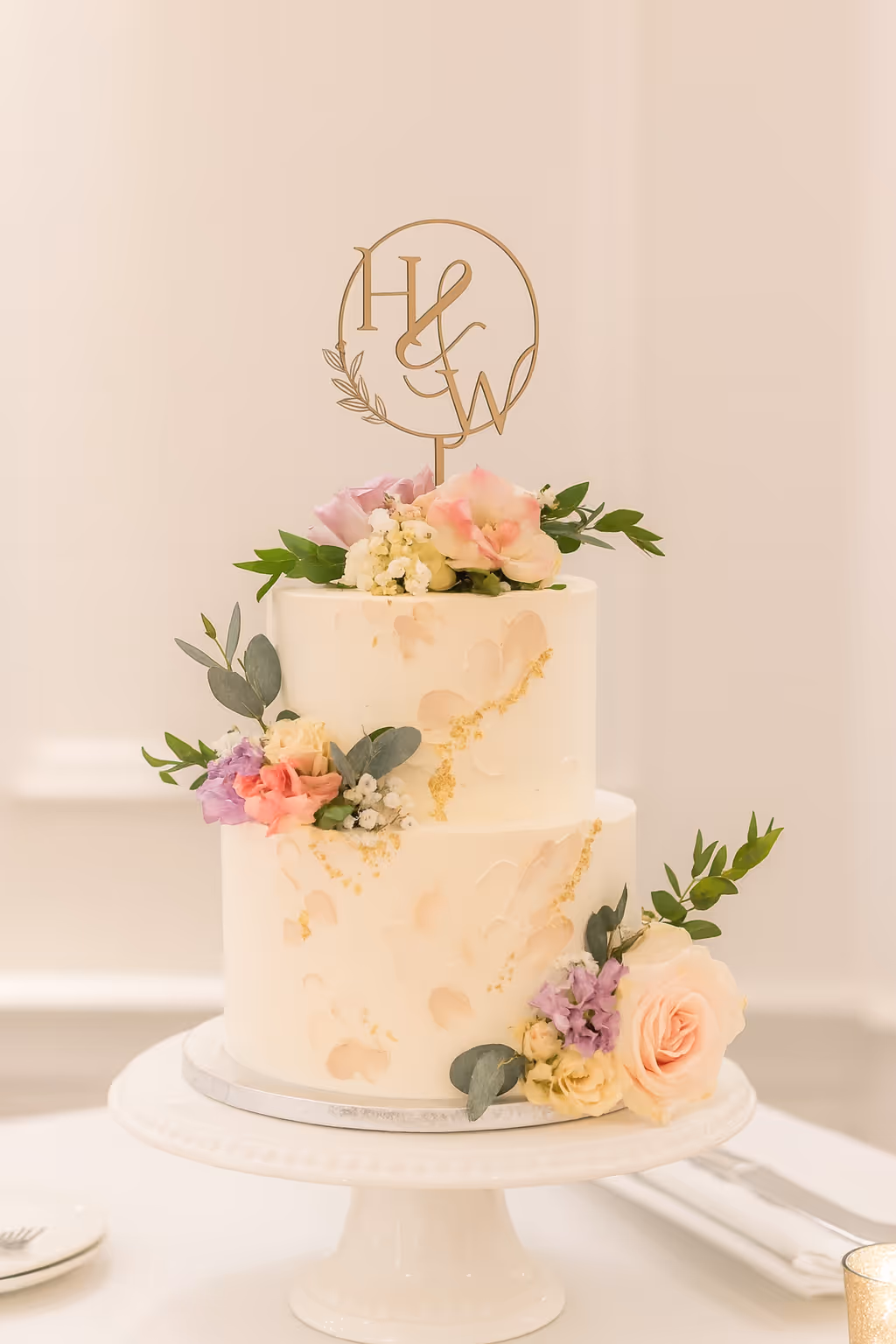 Two-tier cream wedding cake decorated with pastel flowers and gold accents, topped with H&W initials.