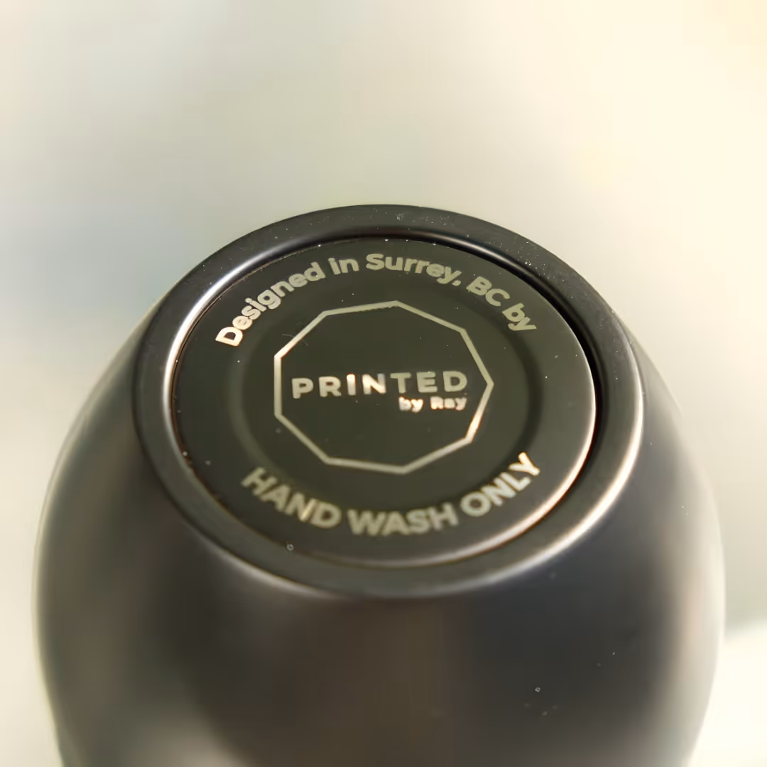 Bottom of a black insulated mug with text: Designed in Surrey BC by Printed by Ray, Hand Wash Only.