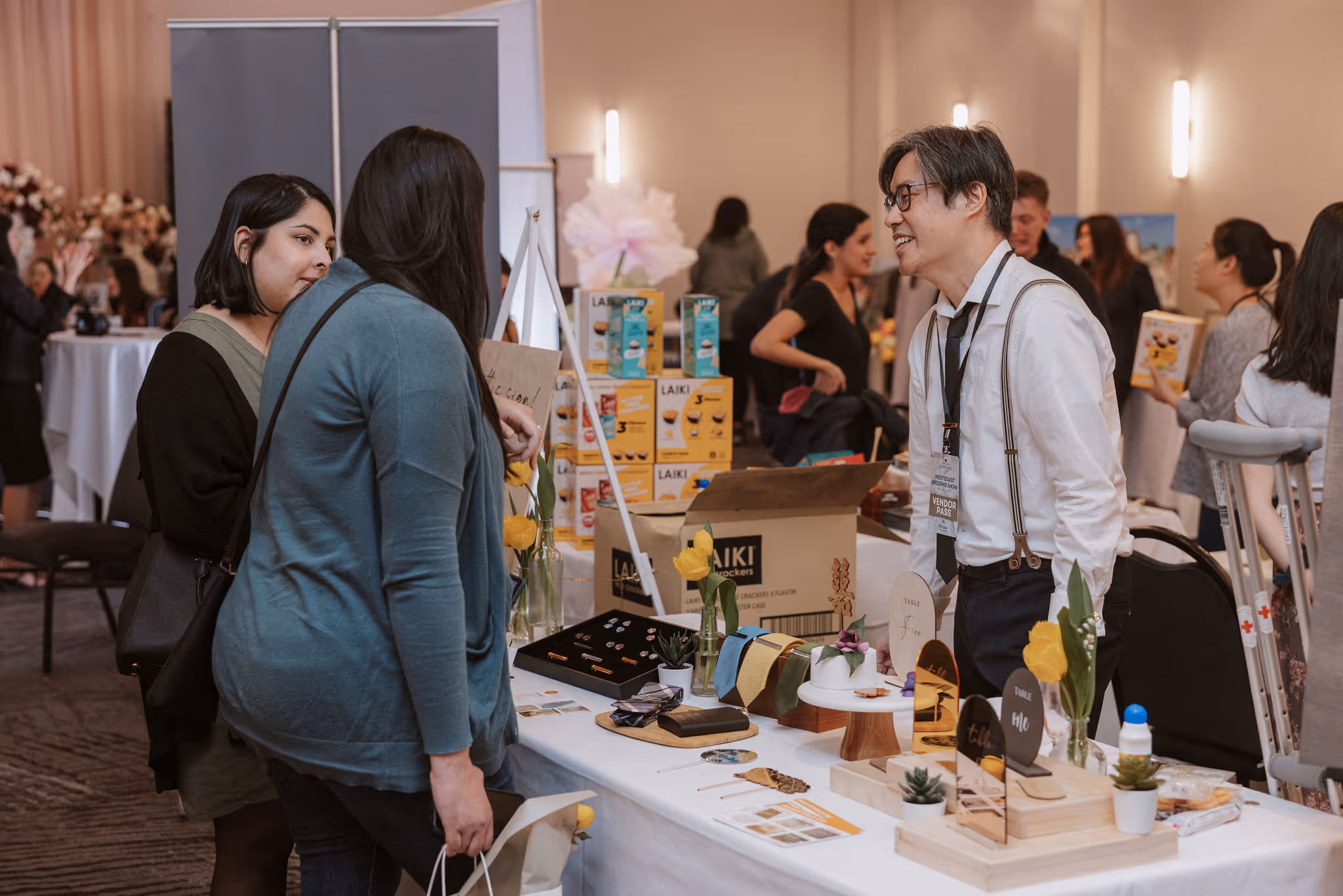 Man wearing vendor pass talks to two women at a table displaying ties and small decor items.