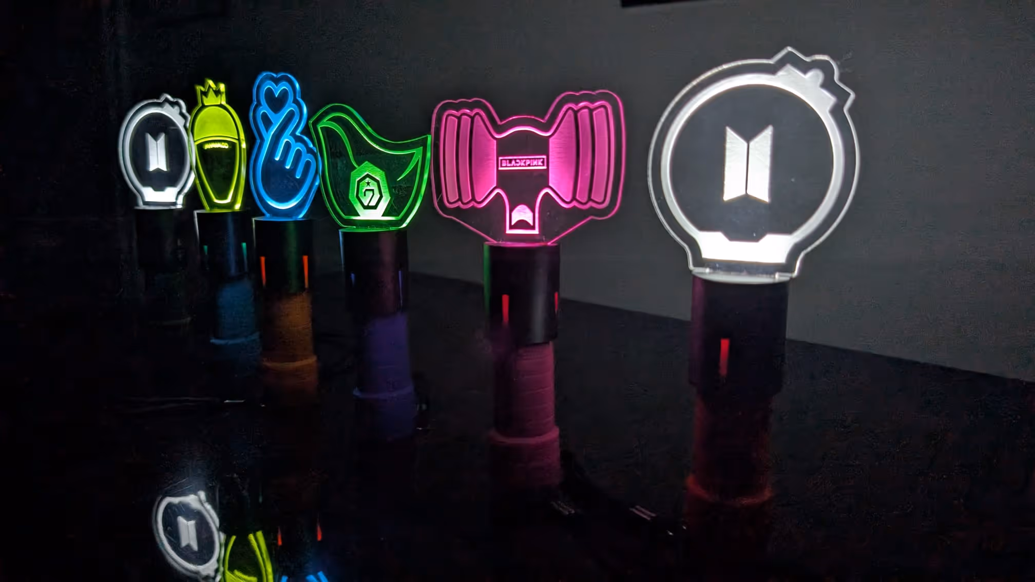 Six colorful K-pop light sticks lit up in a row on a dark reflective surface.