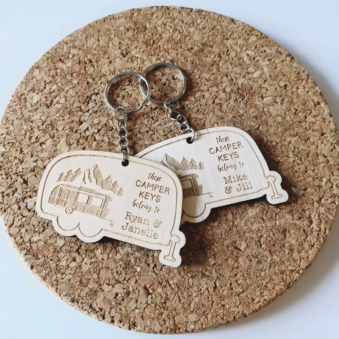 Two wooden camper-shaped keychains with mountain engraving and personalized names on cork mat.