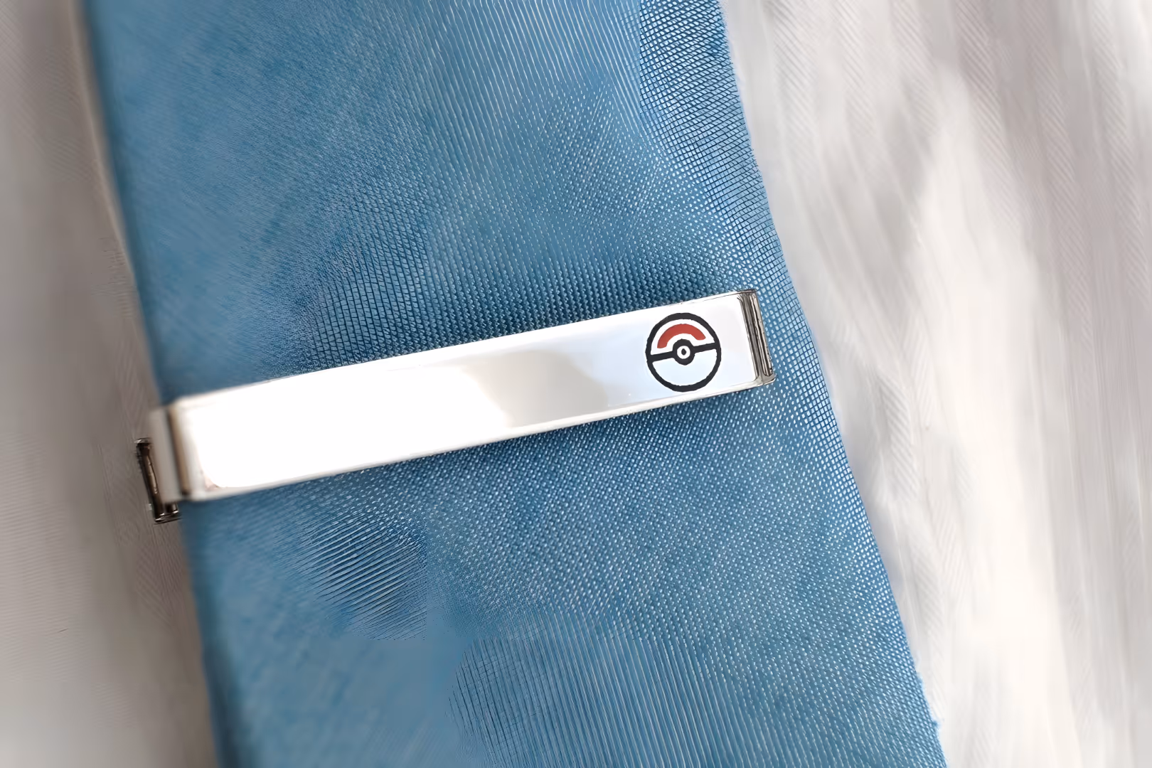 Silver tie clip with a small Poké Ball design resting on a blue textured fabric.