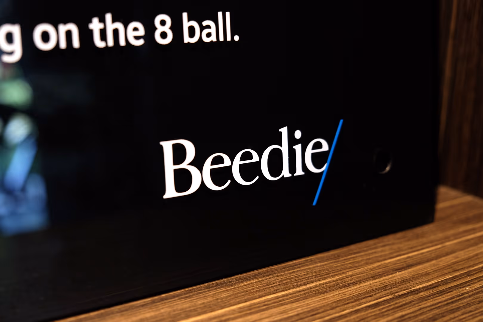 White text 'Beedie' with a blue slash on a black background above a wooden surface.