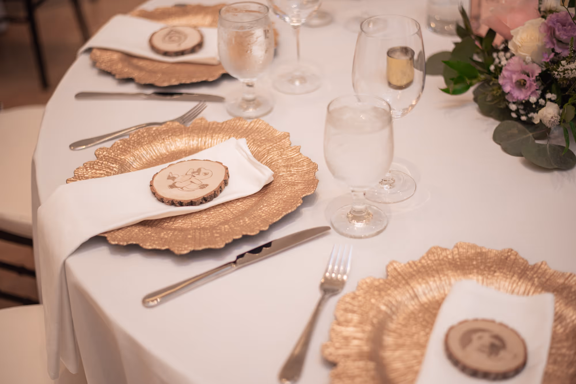 Elegantly set round table with gold charger plates, white napkins, wood slice decor, glasses, and floral centerpiece.
