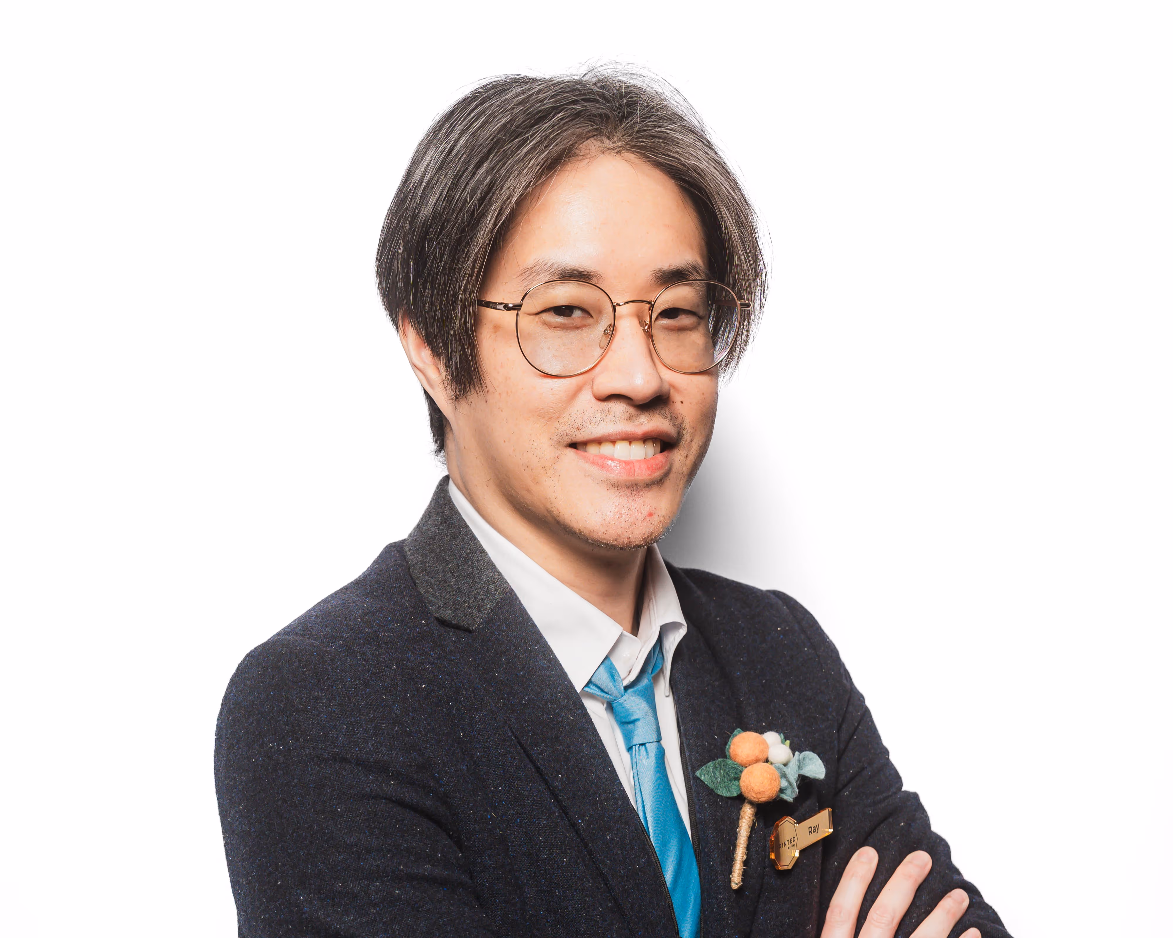 Smiling man with glasses in dark blazer, blue tie, and floral lapel pin posing against white background.