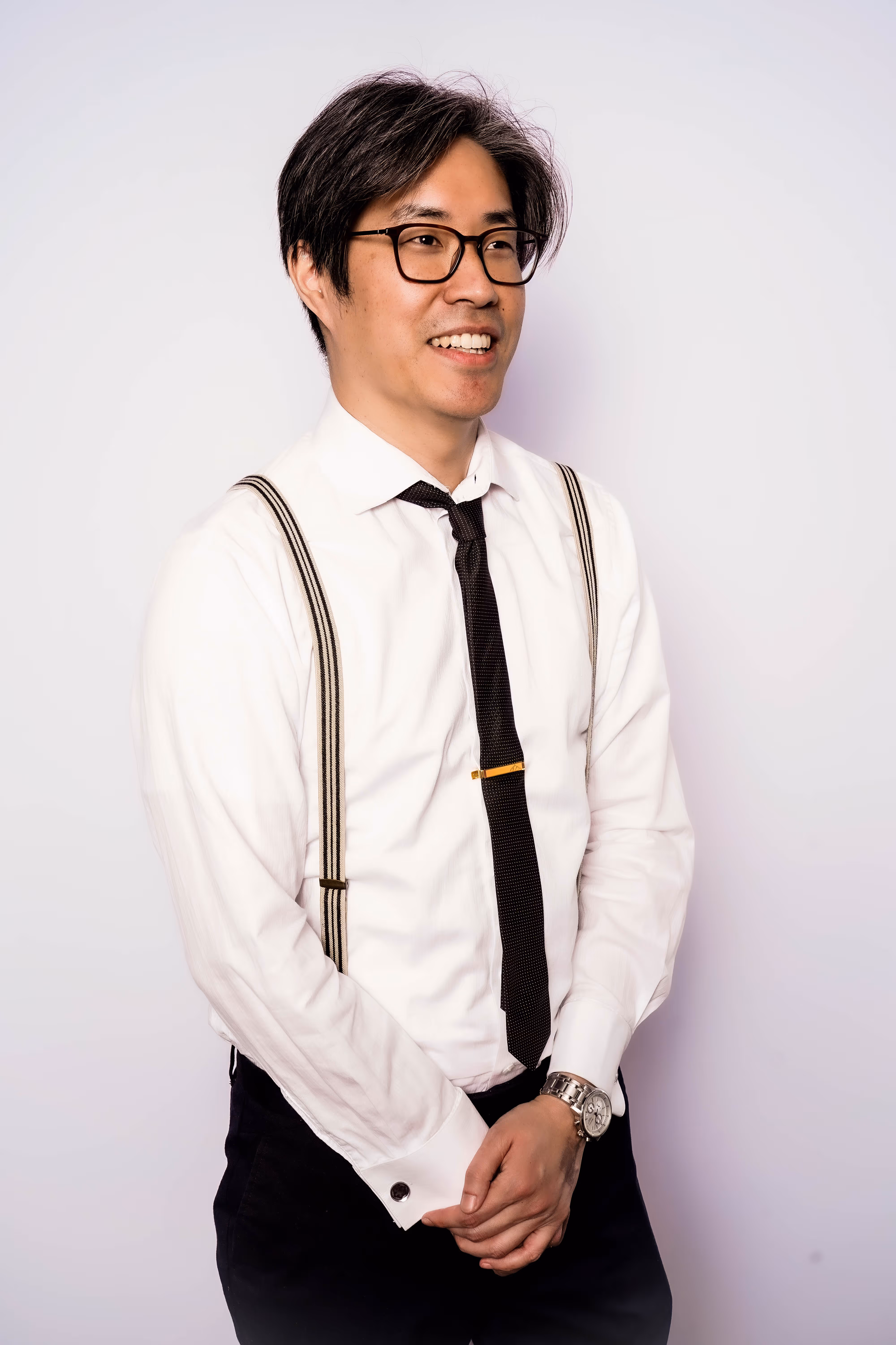 Smiling man with glasses wearing a white shirt, black tie, striped suspenders, and a wristwatch.