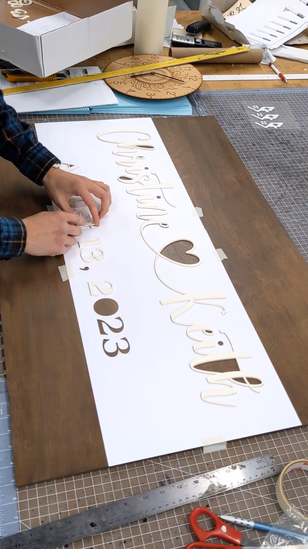 Person arranging cream 3D letters spelling 'Christie Kiritu Aug 13, 2023' on white paper over wood.