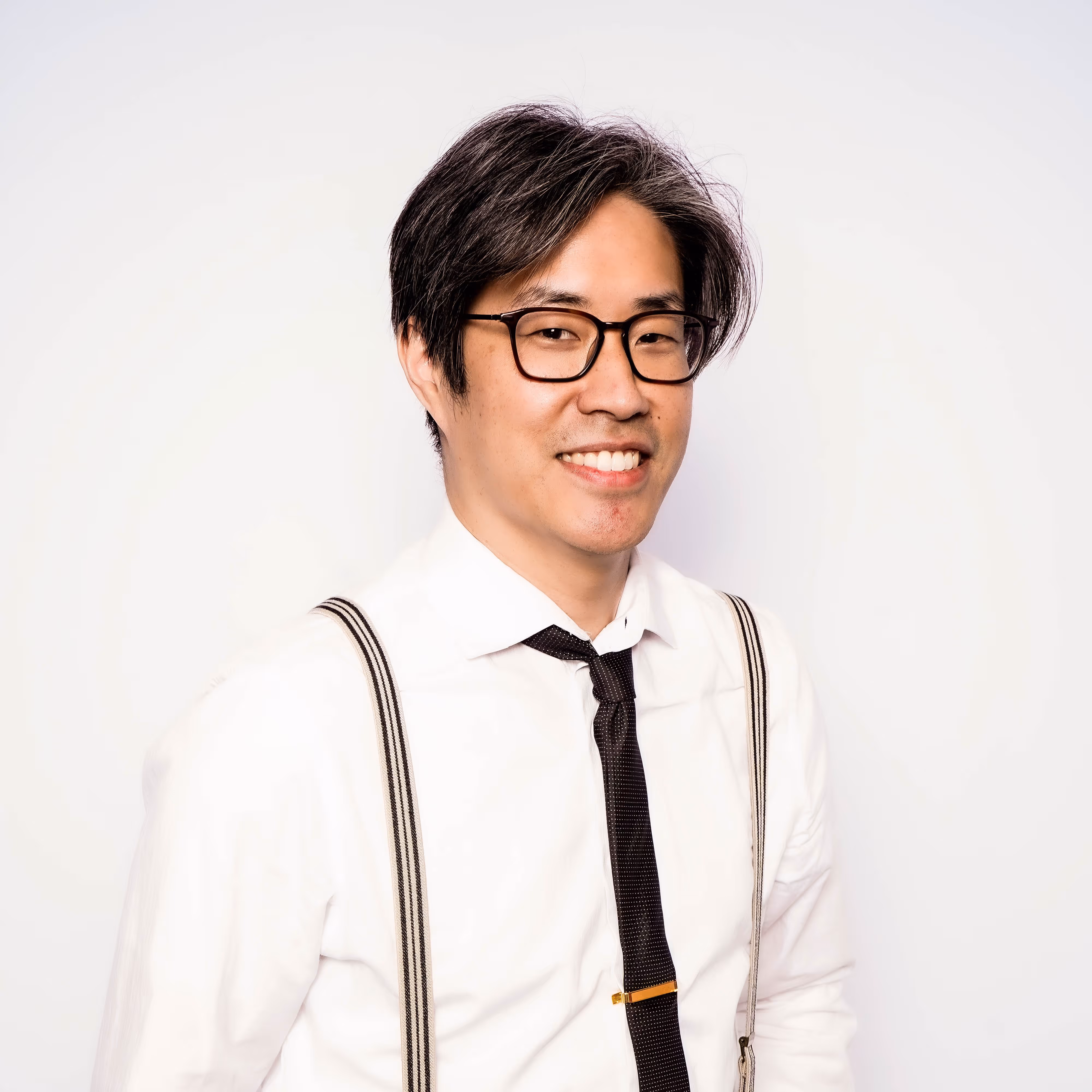 Image of Ray, founder of Printed by Ray. Smiling man with glasses wearing a white shirt, black tie with tie clip, and striped suspenders against a white background.