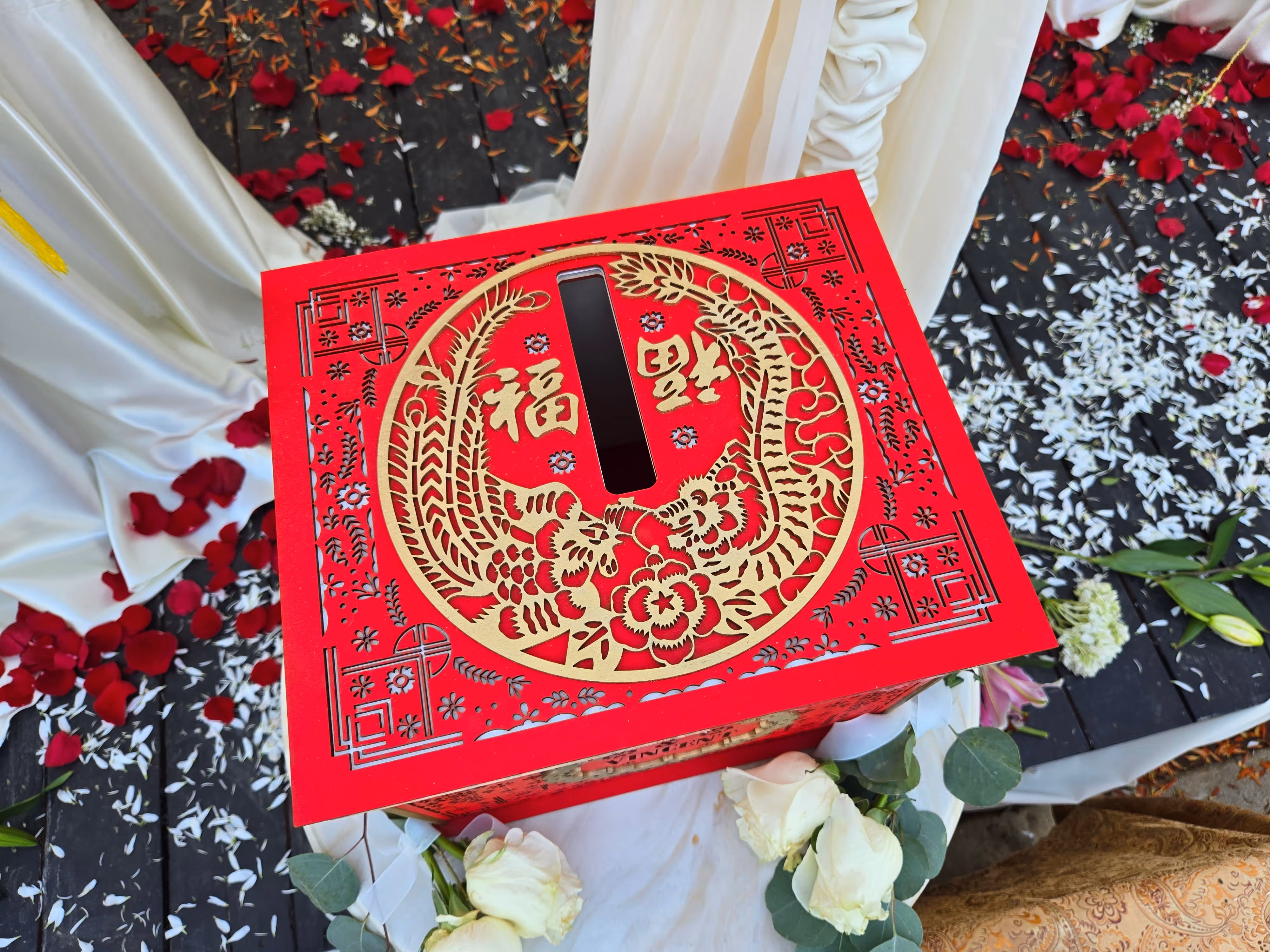 Red and gold Chinese-themed box with floral designs surrounded by white roses and scattered petals.