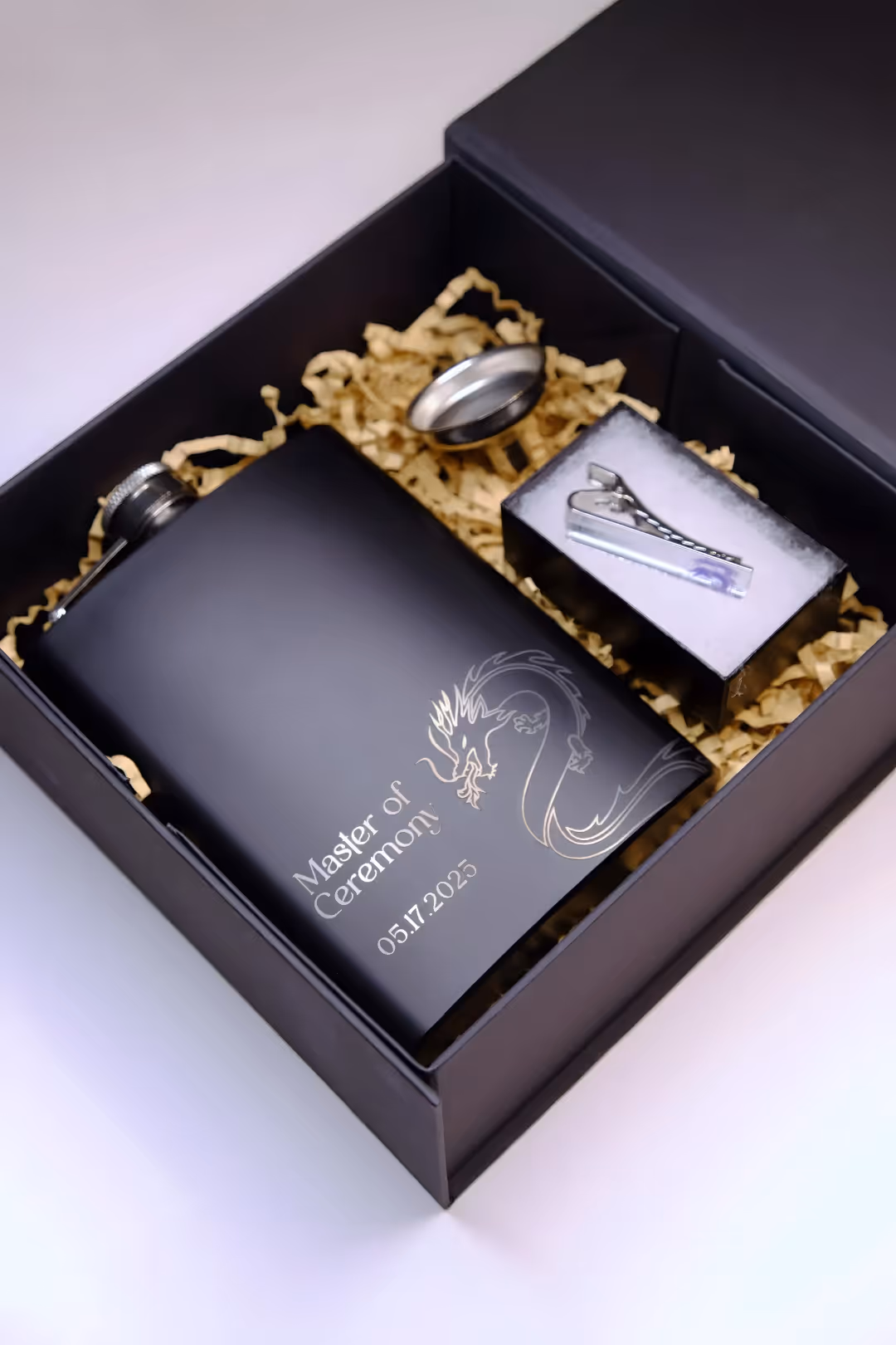 Black gift box with a flask engraved 'Master of Ceremony 05.17.2025,' ring, and tie clip on filler.