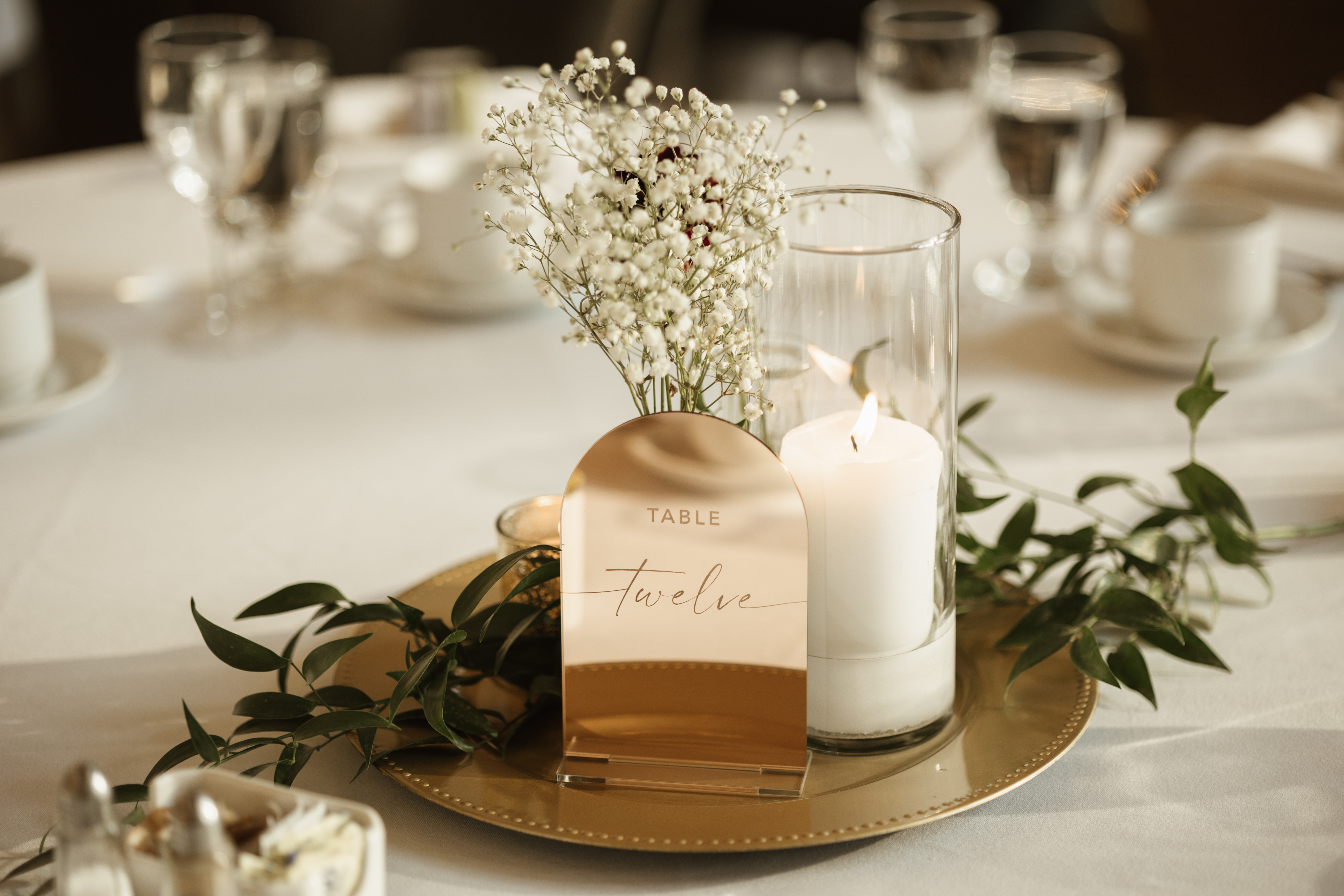 Wedding table centerpiece with lit candle, white flowers, greenery, and a gold table number twelve sign.