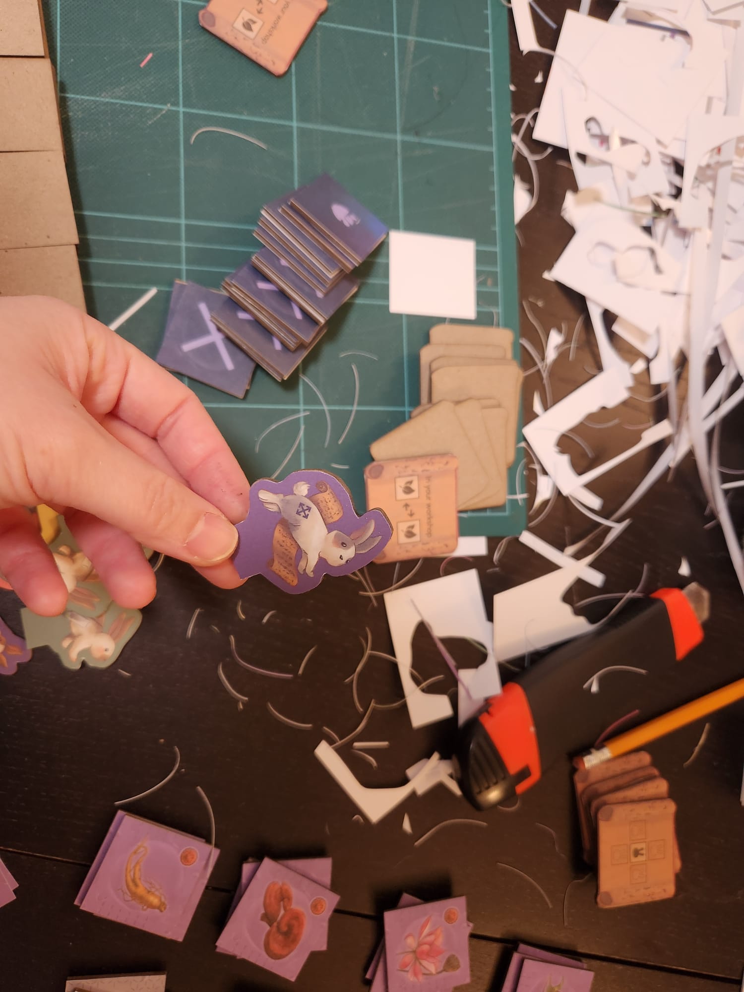Hand holding a purple game piece shaped like a bunny with a scroll, surrounded by board game tokens and crafting tools.