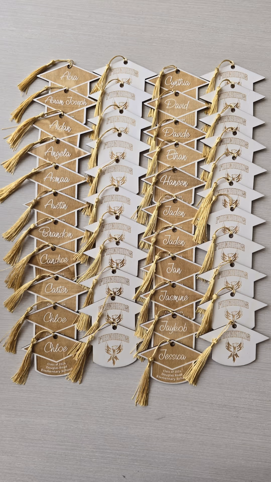 Rows of laser-cut brown and white graduation cap-shaped tags with gold tassels and student names.