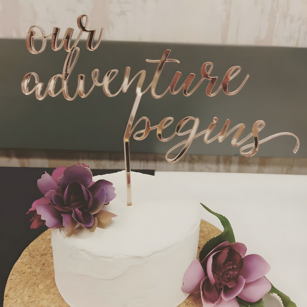 Rose gold acrylic "our adventure begins" cake topper in script lettering, displayed on a white single-tier wedding cake with purple flowers.