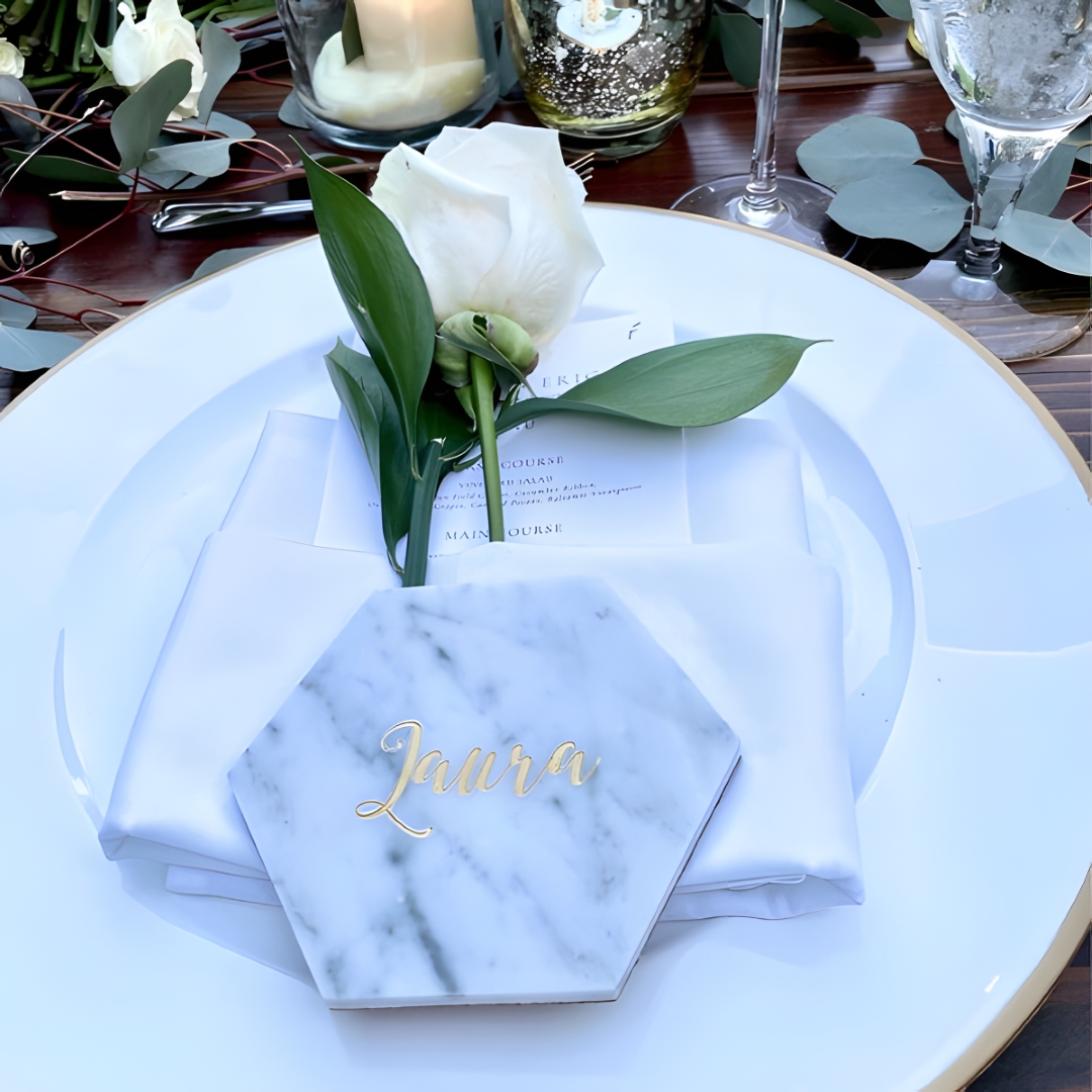Wedding table centrepiece with a frosted hexagonal marble-look acrylic place card reading "Laura" in gold script, on a white plate with a white rose and eucalyptus.