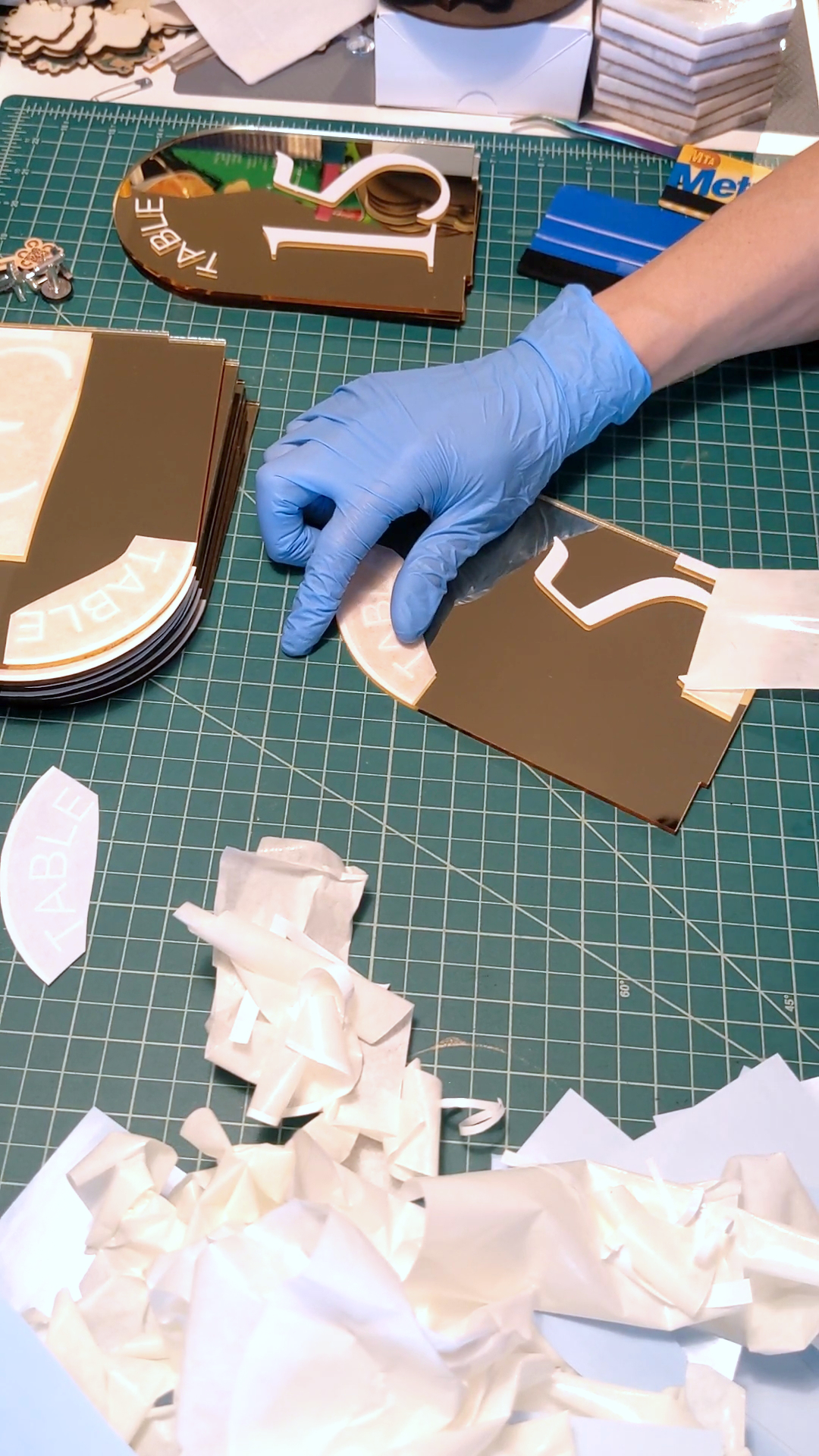 Gloved hand sorting and assembling laser-cut brown acrylic table number pieces on a green cutting mat, surrounded by acrylic offcuts and tools.