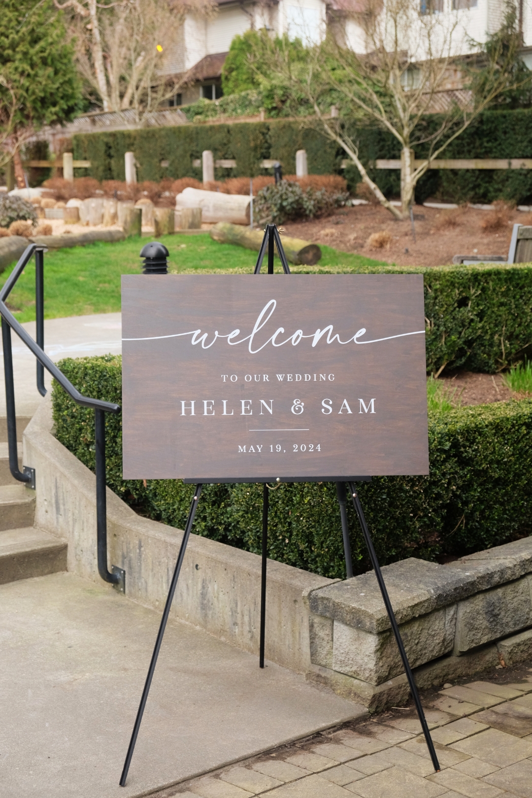 Wood-toned welcome sign reading "Welcome to Our Wedding, Helen & Sam, May 19, 2024," displayed on a black easel outdoors at a garden venue.