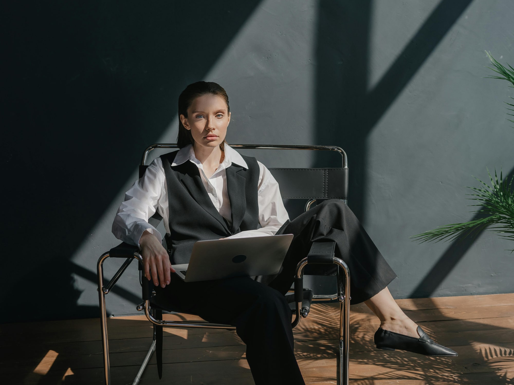 Woman in black vest and white shirt sitting on a chair with a laptop on her lap in a room with dark walls.