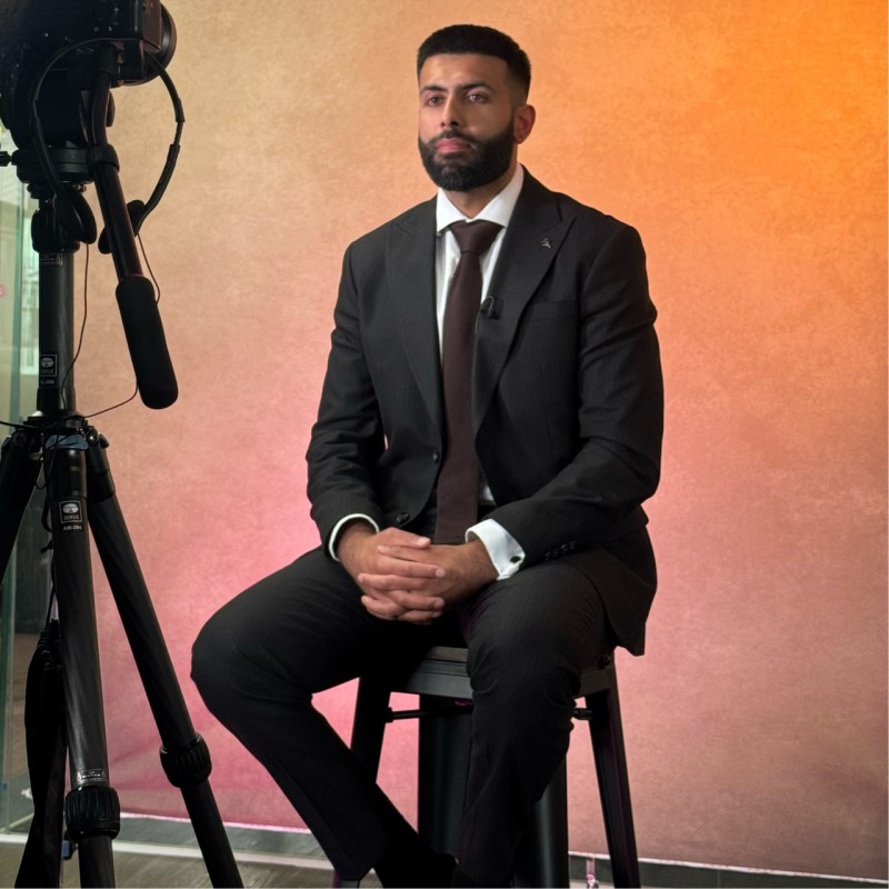 Man in a black suit sitting on a stool with hands clasped, studio camera setup on the left.