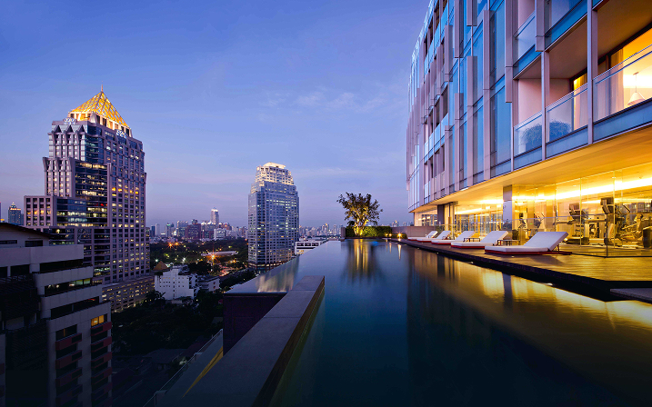 Infinity pool on rooftop next to modern building overlooking cityscape at dusk with lounge chairs.