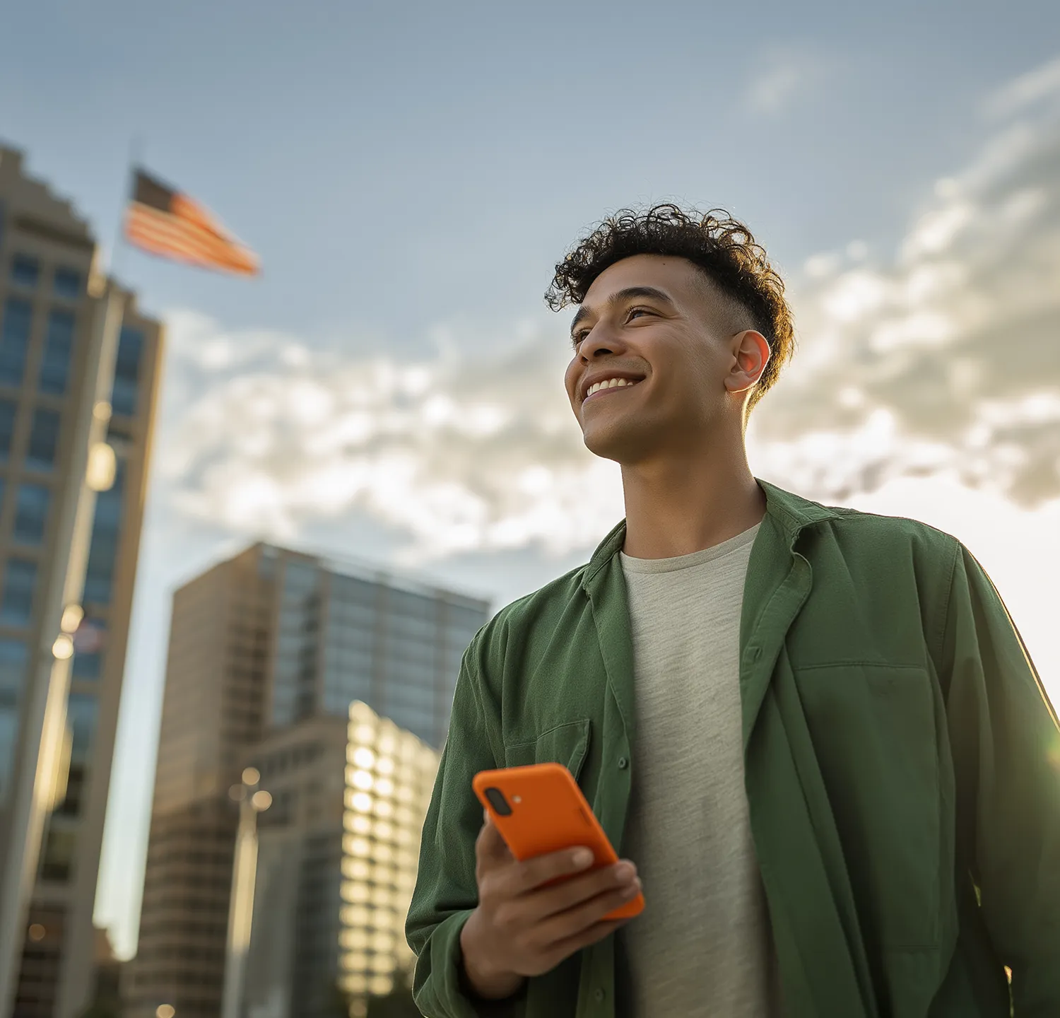 Joven sonriente con camisa verde y teléfono naranja en la mano, con edificios y bandera al fondo.