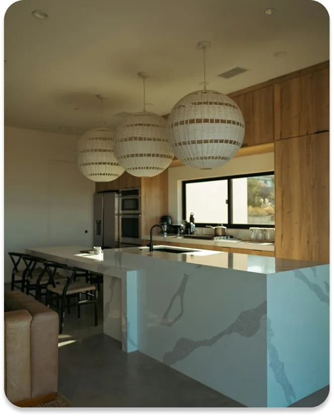 Modern kitchen with a marble island, wooden cabinets, black chairs, and three large hanging lights.