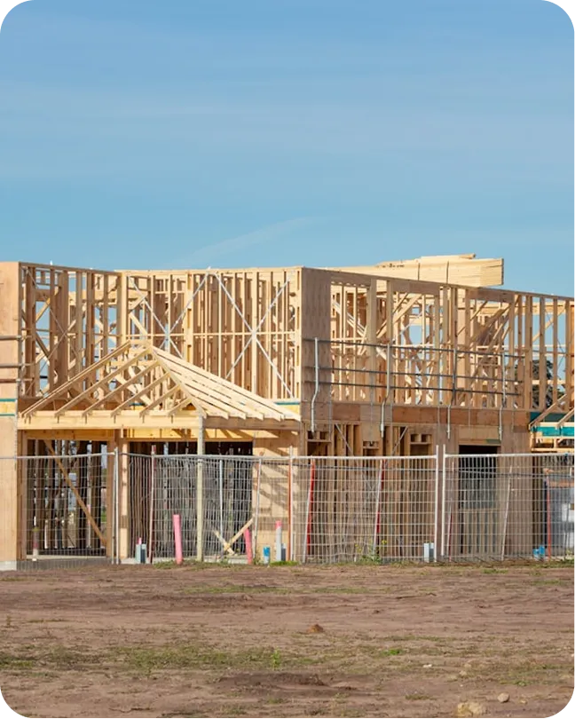 Wooden framework of a two-story house under construction on a clear day with fenced perimeter.
