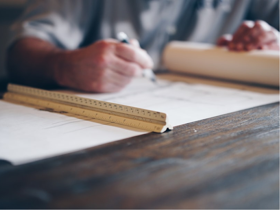 Person drawing on paper with a pen and wooden ruler on a dark wooden table.
